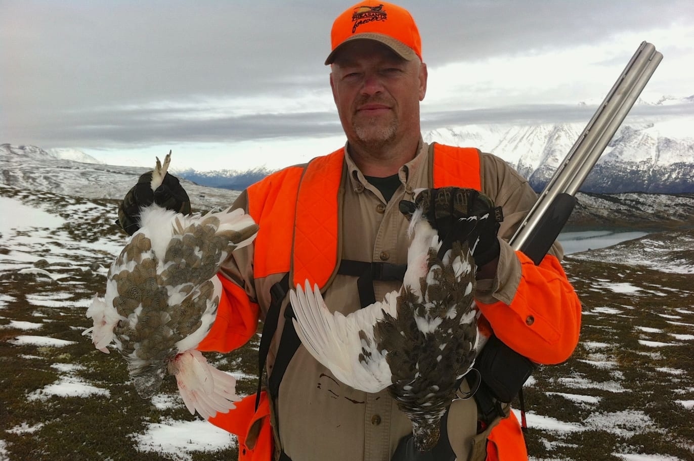 A hunter poses with two ptarmigans in front of a snowy Alaskan peak.