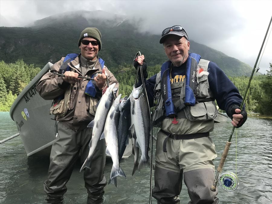 Misty mountain peaks provide a picturesque backdrop for a day of fishing. Two fishermen hold up salmon caught on a fly rod.