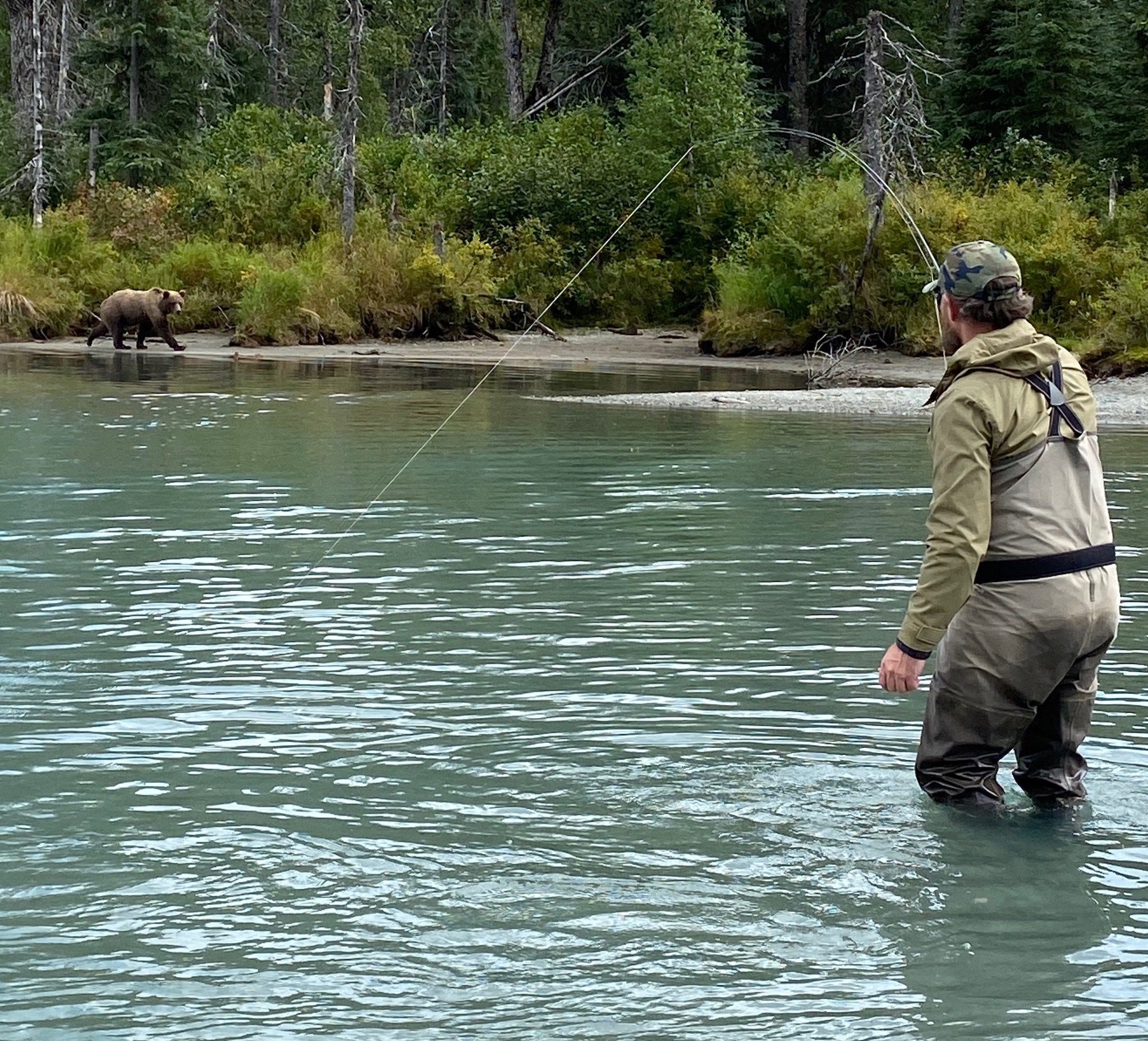 A fly fisherman fights a fish in turquoise water while a small bear looks on.